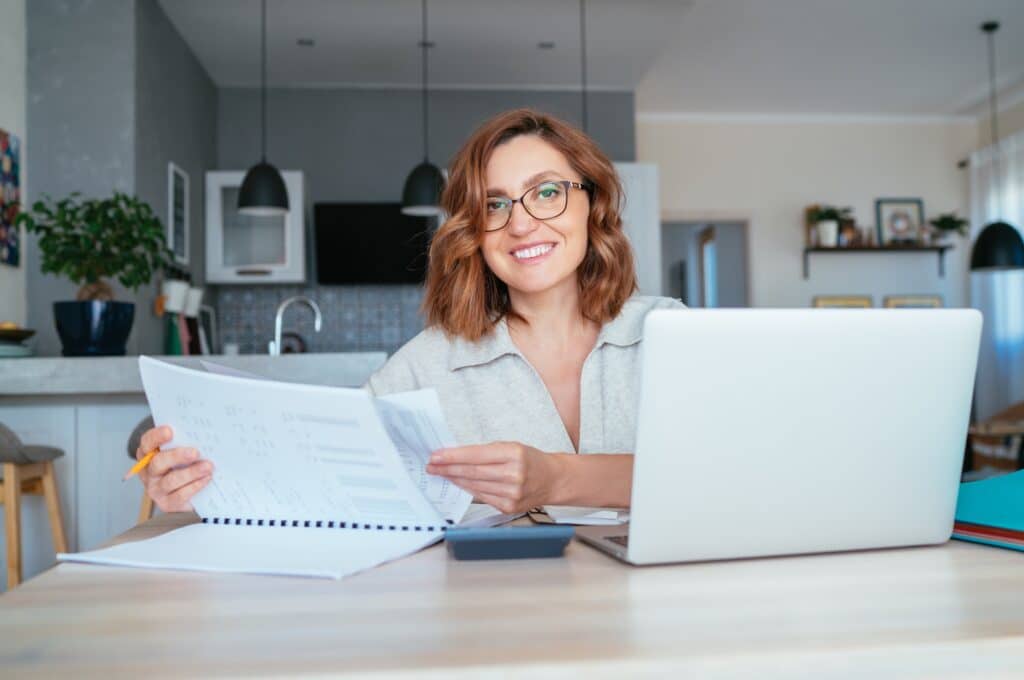 Beautiful middle aged woman in glasses smiling at camera. She does Quarterly financial reports at the home office using modern laptop. Small business, home finance, money savings concept image.