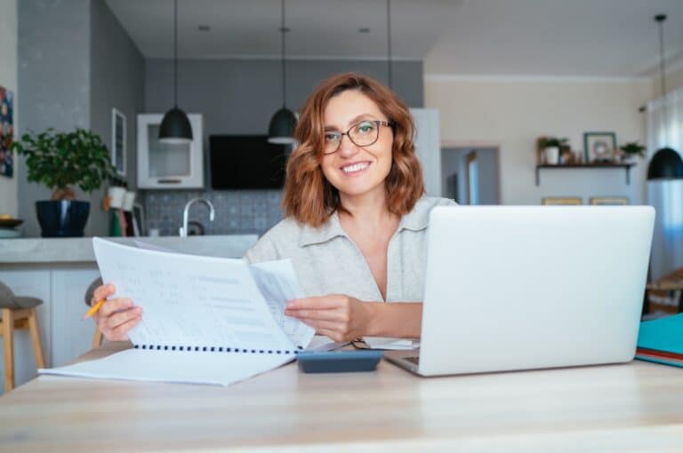 Beautiful middle aged woman in glasses smiling at camera. She does Quarterly financial reports at the home office using modern laptop. Small business, home finance, money savings concept image.